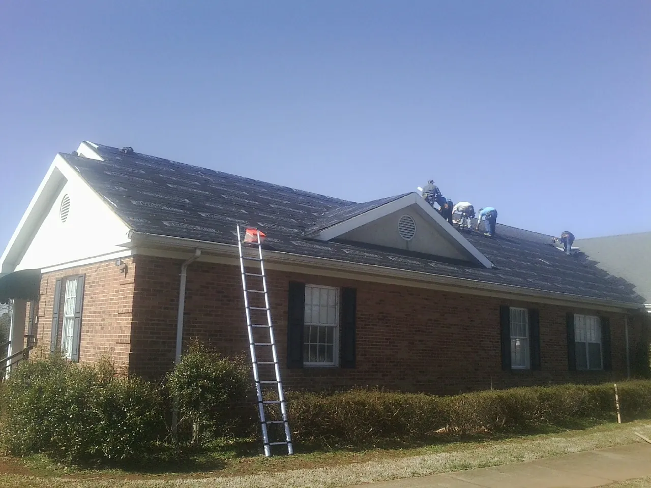Crew tearing shingles off a brick ranch roof with a ladder set — Emergency Roof Repair work by our crew in Cherry Valley